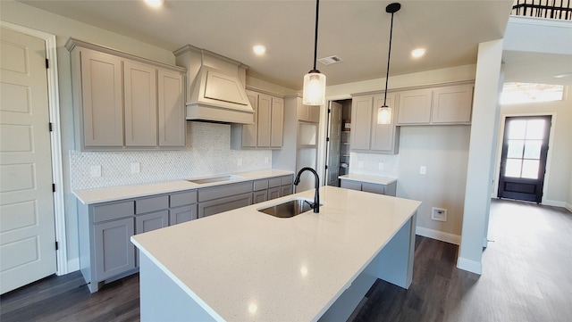 kitchen with sink, custom exhaust hood, hanging light fixtures, dark hardwood / wood-style flooring, and a kitchen island with sink