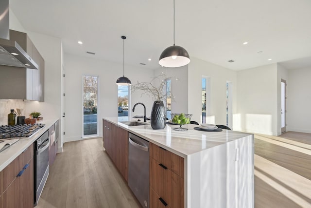 kitchen with sink, a large island with sink, stainless steel appliances, light wood-type flooring, and wall chimney exhaust hood