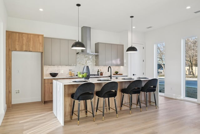 kitchen featuring pendant lighting, backsplash, a center island with sink, wall chimney exhaust hood, and light wood-type flooring