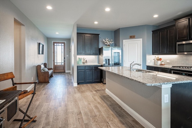 kitchen with a kitchen island with sink, a sink, light wood-style floors, appliances with stainless steel finishes, and decorative backsplash