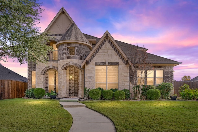 french country inspired facade featuring stone siding, a front yard, a balcony, and fence