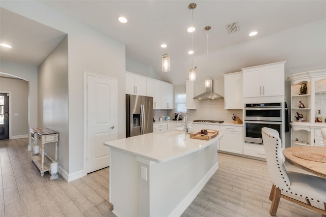 kitchen featuring pendant lighting, wall chimney range hood, appliances with stainless steel finishes, an island with sink, and white cabinets