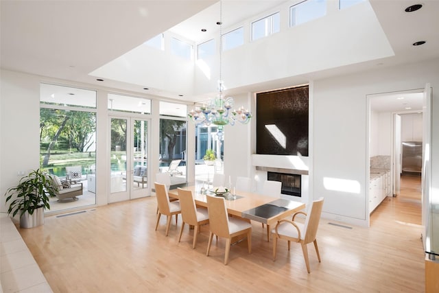 dining area with a towering ceiling, a notable chandelier, a fireplace, light hardwood / wood-style floors, and french doors