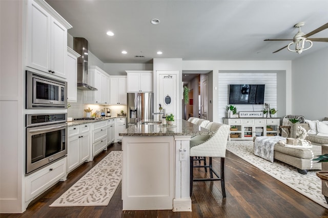 kitchen featuring appliances with stainless steel finishes, light stone counters, white cabinets, a center island with sink, and wall chimney exhaust hood