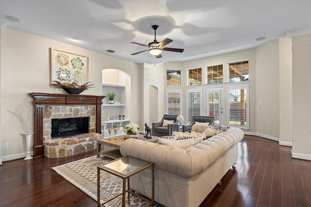 living room featuring ceiling fan, built in shelves, a stone fireplace, and dark wood-type flooring