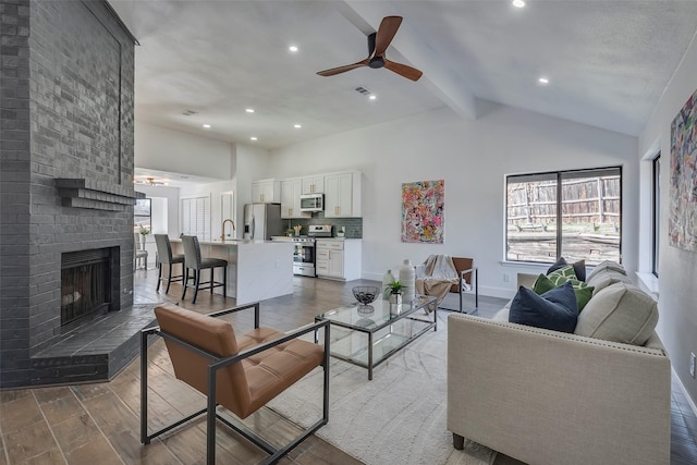 living room featuring high vaulted ceiling, beamed ceiling, sink, ceiling fan, and a brick fireplace