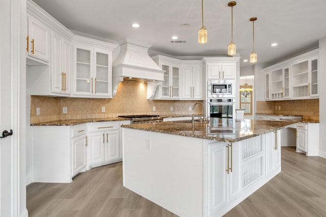 kitchen with appliances with stainless steel finishes, white cabinets, dark stone counters, custom range hood, and a center island with sink