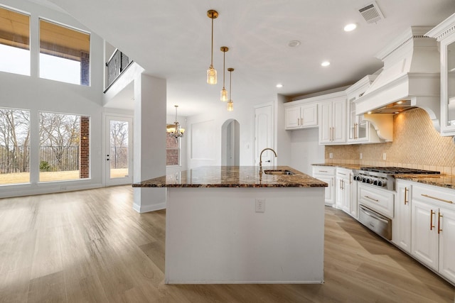 kitchen featuring an island with sink, custom range hood, white cabinets, and dark stone counters