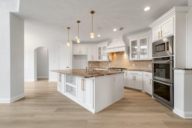 kitchen featuring a kitchen island with sink, white cabinetry, dark stone countertops, stainless steel appliances, and custom exhaust hood