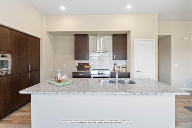 kitchen with an island with sink, sink, stainless steel appliances, light stone countertops, and wall chimney exhaust hood