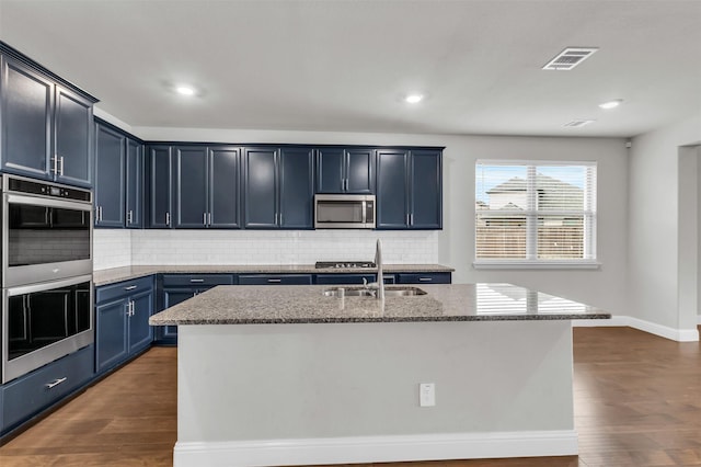 kitchen with appliances with stainless steel finishes, an island with sink, sink, dark wood-type flooring, and blue cabinetry