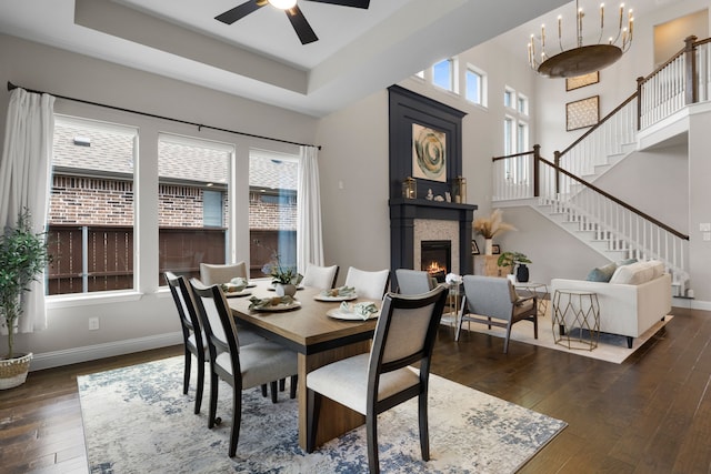 dining space featuring a large fireplace, dark hardwood / wood-style floors, a raised ceiling, and ceiling fan with notable chandelier