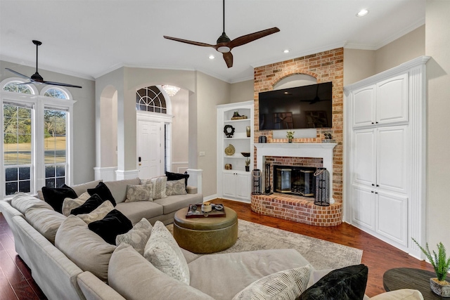 living room with dark wood-type flooring, ceiling fan, ornamental molding, and a brick fireplace