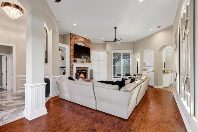 living room featuring built in features, dark hardwood / wood-style floors, ornamental molding, a brick fireplace, and ceiling fan with notable chandelier