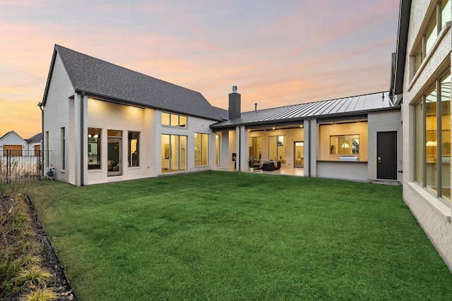 back of property at dusk featuring a patio, a standing seam roof, a yard, a chimney, and metal roof