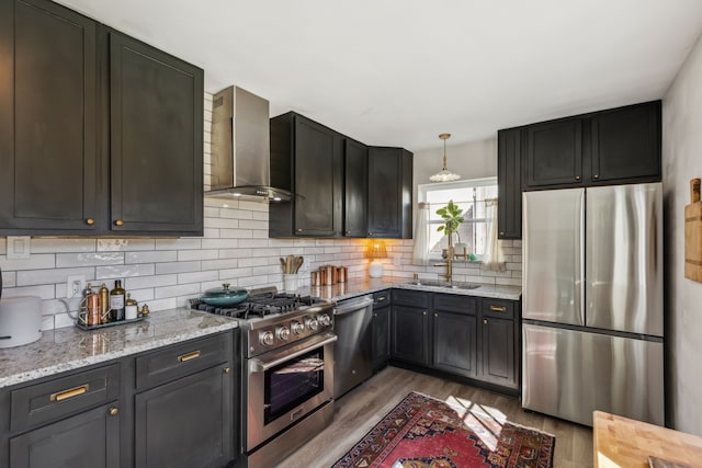 kitchen with wall chimney exhaust hood, light stone counters, hanging light fixtures, stainless steel appliances, and light hardwood / wood-style floors