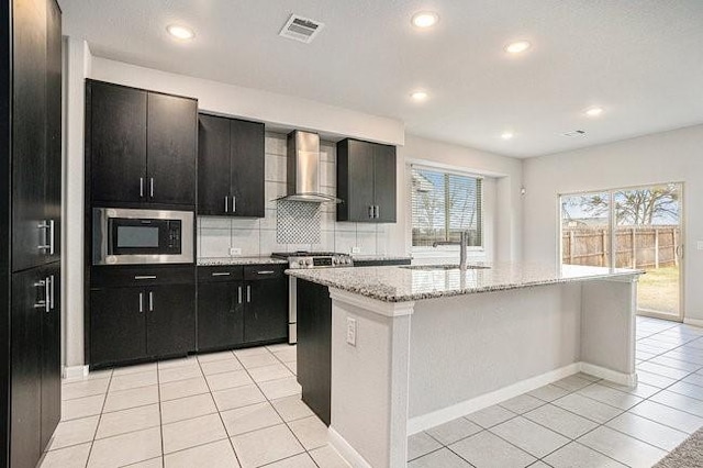 kitchen featuring built in microwave, stainless steel gas range, wall chimney range hood, a kitchen island with sink, and backsplash