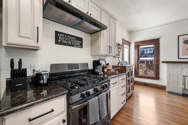 kitchen with dark stone countertops, white cabinets, a brick fireplace, stainless steel range with gas stovetop, and light hardwood / wood-style flooring