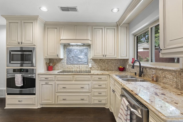 kitchen with sink, custom exhaust hood, stainless steel appliances, light stone countertops, and cream cabinetry