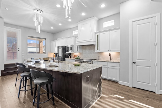 kitchen featuring range, white cabinets, an island with sink, stainless steel microwave, and custom exhaust hood