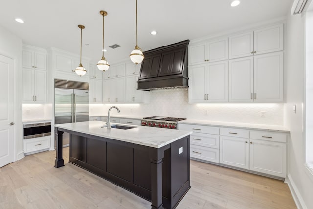 kitchen featuring sink, premium range hood, built in refrigerator, white cabinets, and a center island with sink