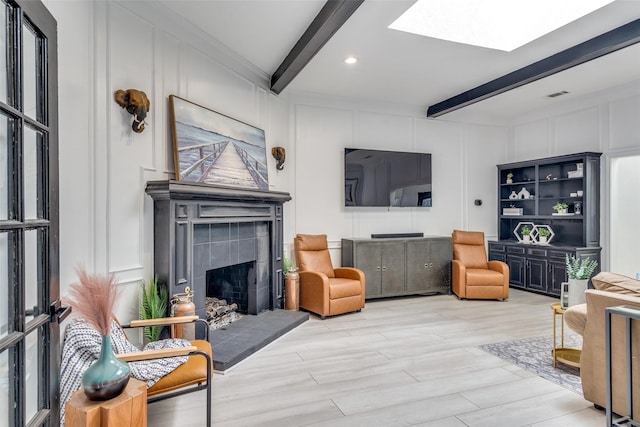 living room featuring beamed ceiling, a skylight, a tiled fireplace, and light wood-type flooring