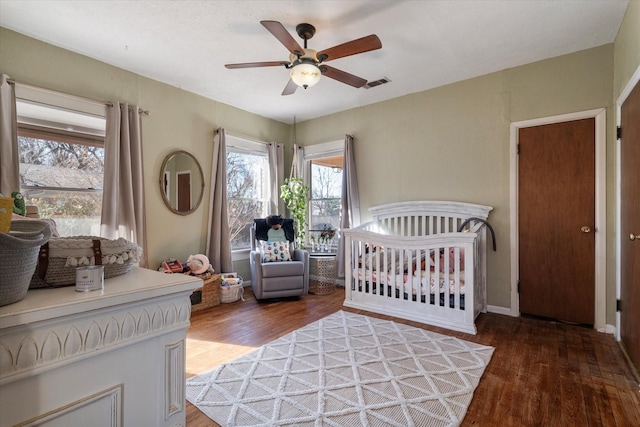 bedroom with a nursery area, dark wood-type flooring, and ceiling fan