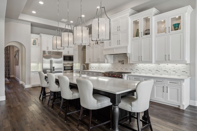 kitchen with built in appliances, pendant lighting, an island with sink, and white cabinets