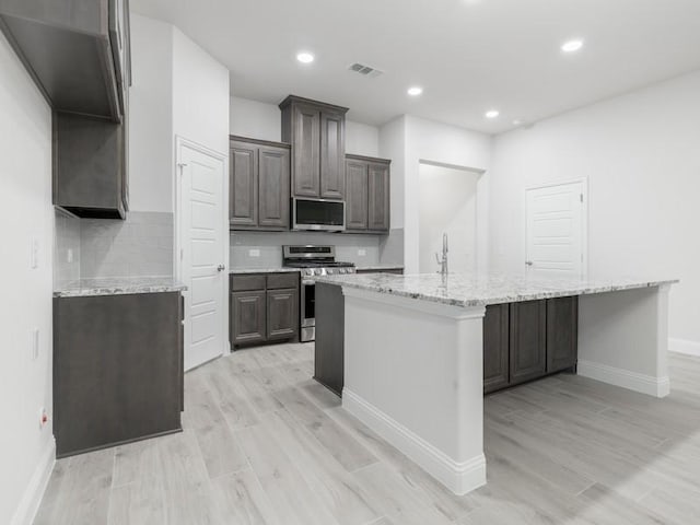 kitchen featuring appliances with stainless steel finishes, light stone countertops, a kitchen island with sink, and light wood-type flooring