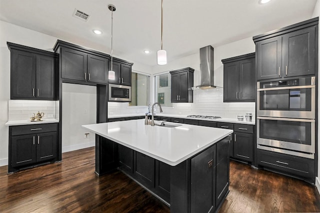 kitchen with sink, stainless steel appliances, a center island with sink, decorative light fixtures, and wall chimney exhaust hood