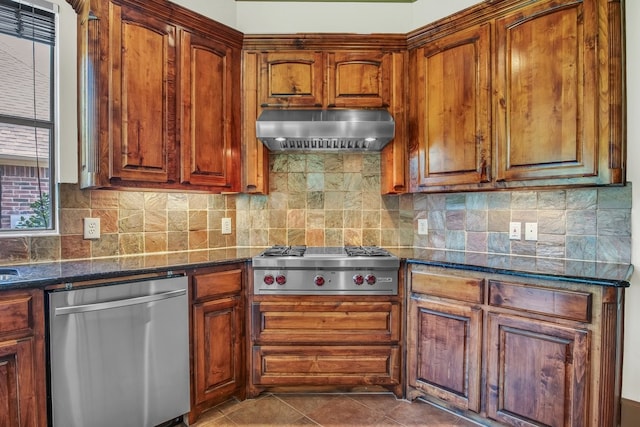 kitchen featuring dark stone countertops, range hood, stainless steel appliances, dark tile patterned flooring, and decorative backsplash