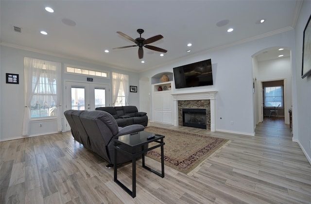 living room featuring a stone fireplace, ornamental molding, ceiling fan, light wood-type flooring, and french doors