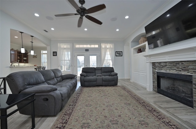 living room featuring ceiling fan, ornamental molding, light hardwood / wood-style floors, a stone fireplace, and french doors