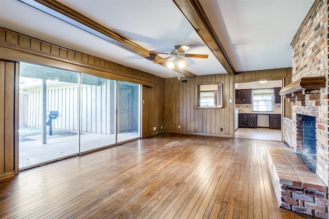 unfurnished living room featuring a fireplace, wood walls, beam ceiling, ceiling fan, and light wood-type flooring