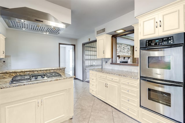 kitchen featuring light tile patterned flooring, appliances with stainless steel finishes, backsplash, light stone counters, and wall chimney range hood