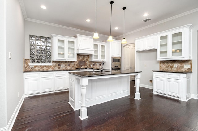 kitchen with glass insert cabinets, dark countertops, premium range hood, and an island with sink