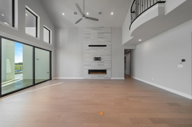 unfurnished living room with ceiling fan, a towering ceiling, a tiled fireplace, and light hardwood / wood-style floors