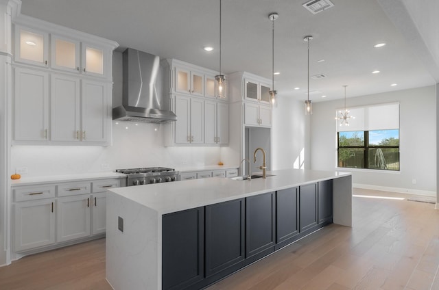 kitchen featuring wall chimney range hood, sink, white cabinetry, a kitchen island with sink, and hanging light fixtures