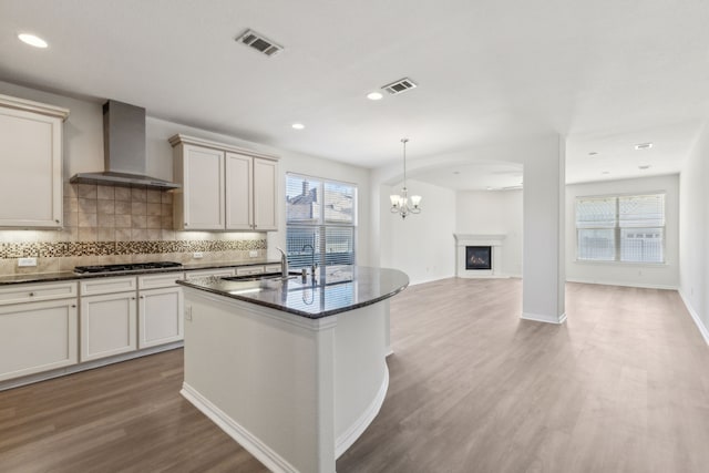 kitchen featuring decorative light fixtures, stainless steel gas stovetop, hardwood / wood-style flooring, a center island with sink, and wall chimney exhaust hood