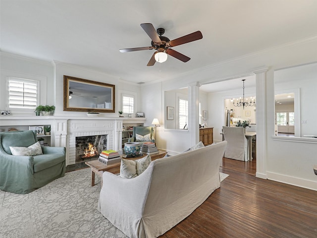 living room with dark wood-type flooring, decorative columns, ornamental molding, a brick fireplace, and ceiling fan with notable chandelier