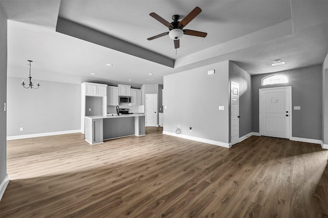 unfurnished living room featuring a raised ceiling, a sink, wood finished floors, baseboards, and ceiling fan with notable chandelier