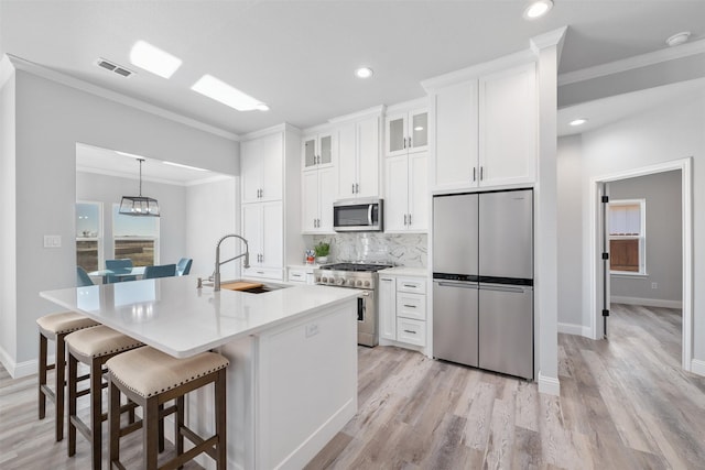 kitchen with sink, a kitchen island with sink, stainless steel appliances, ornamental molding, and white cabinets