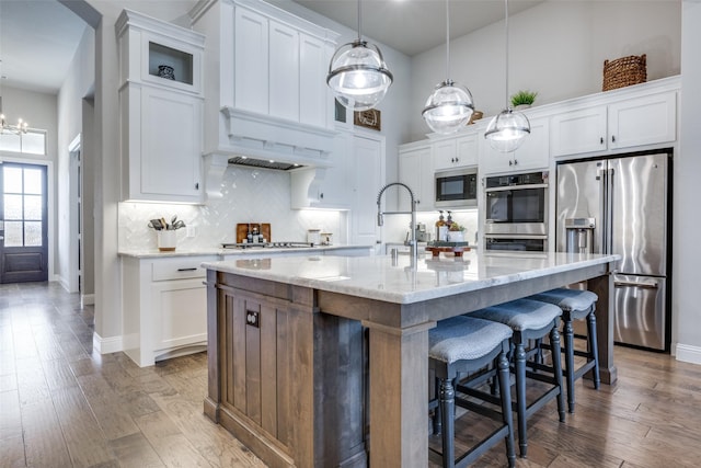 kitchen featuring a kitchen island with sink, decorative light fixtures, stainless steel appliances, and white cabinetry