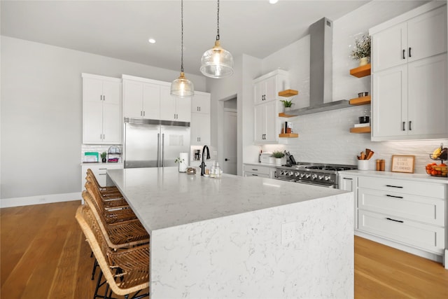 kitchen with wall chimney range hood, a kitchen island with sink, white cabinetry, high quality appliances, and a kitchen bar