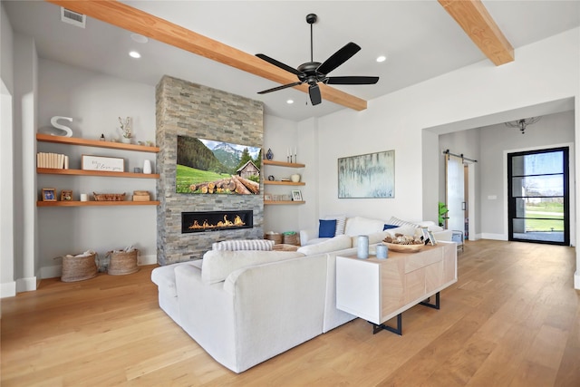 living room with a barn door, beamed ceiling, ceiling fan, a fireplace, and light hardwood / wood-style floors