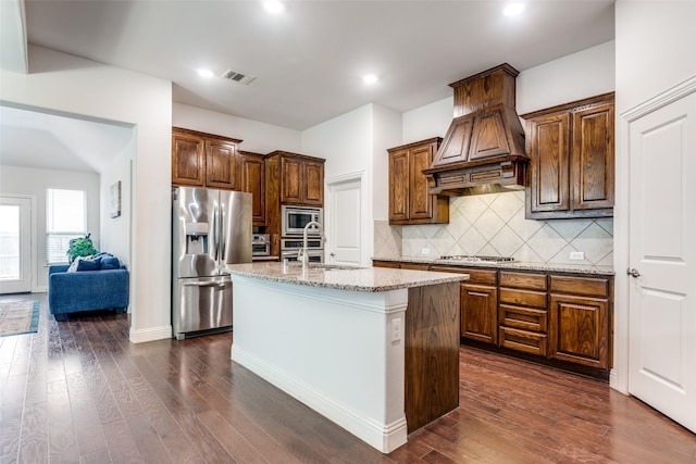 kitchen with appliances with stainless steel finishes, a center island with sink, dark hardwood / wood-style flooring, decorative backsplash, and custom exhaust hood