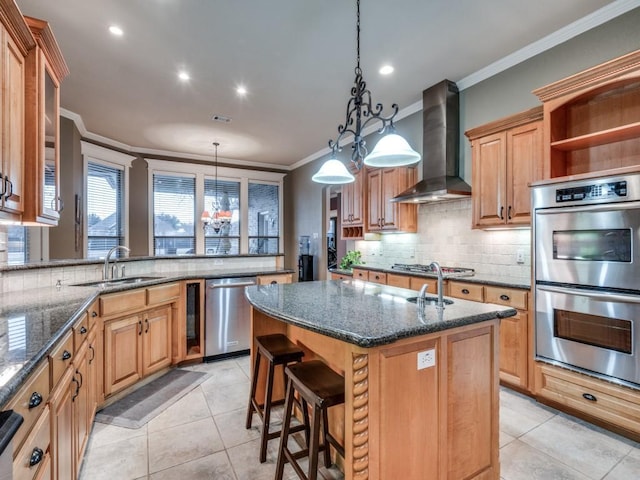 kitchen with appliances with stainless steel finishes, an island with sink, sink, hanging light fixtures, and wall chimney exhaust hood