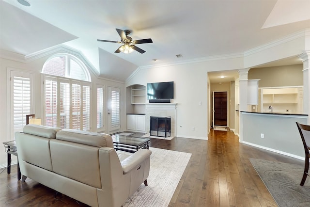 living room with dark wood-type flooring, ceiling fan, a tiled fireplace, built in shelves, and ornate columns