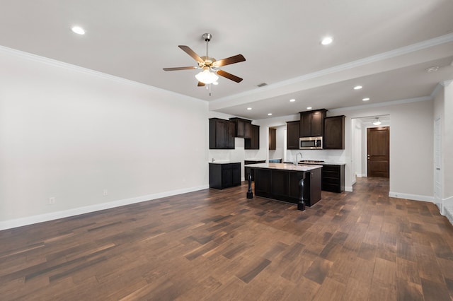 kitchen with dark wood-type flooring, dark brown cabinetry, ornamental molding, an island with sink, and ceiling fan