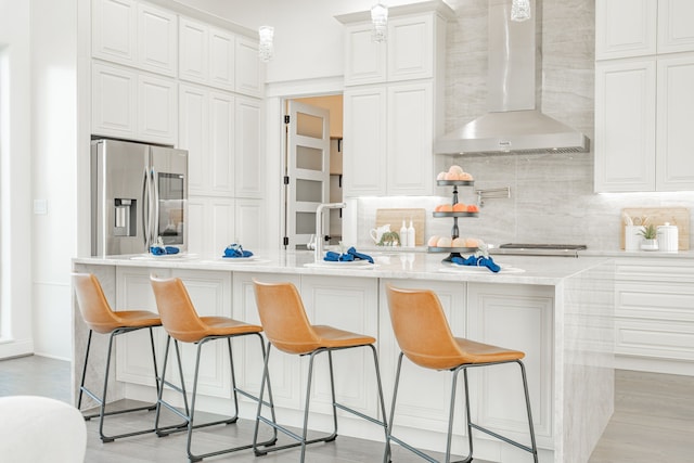 kitchen with wall chimney range hood, stainless steel fridge, white cabinets, and a center island with sink